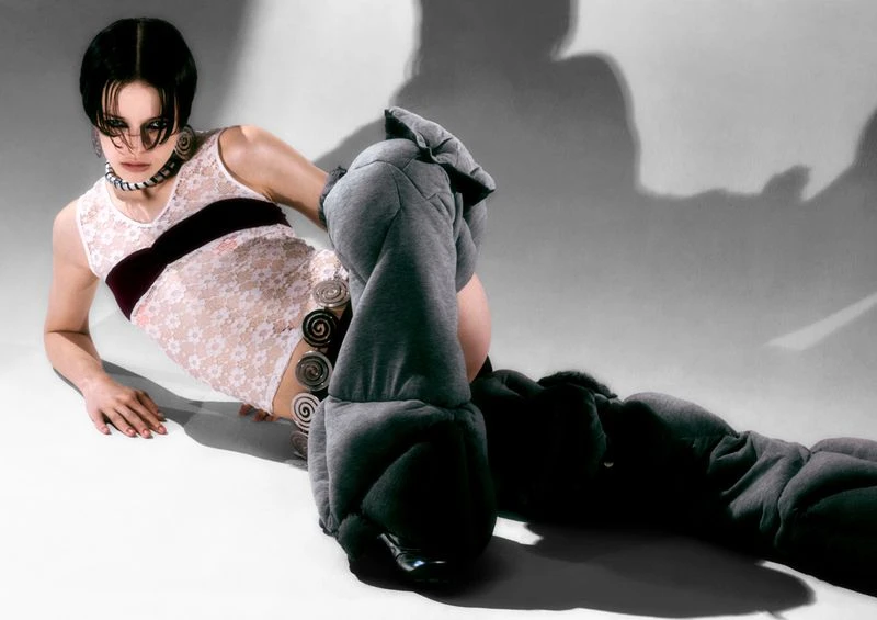Female model in a white lace bodysuit with dark brown trim and oversized grey leg coverings in a modern fashion studio.