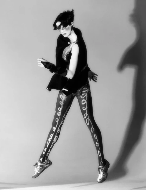 Black and white studio fashion shot of a woman in patterned tights jumping, wearing a jacket and jewelry against a neutral backdrop.