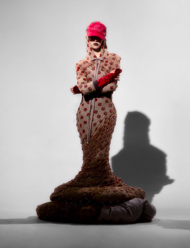 A fashionably dressed person poses in a couture gown, cap, and gloves, creating a unique and visually striking full-length studio shot.