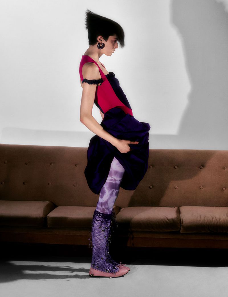 A woman in unique fashion stands in a photo studio with a brown couch, wearing purple patterned tights and dark purple skirt.