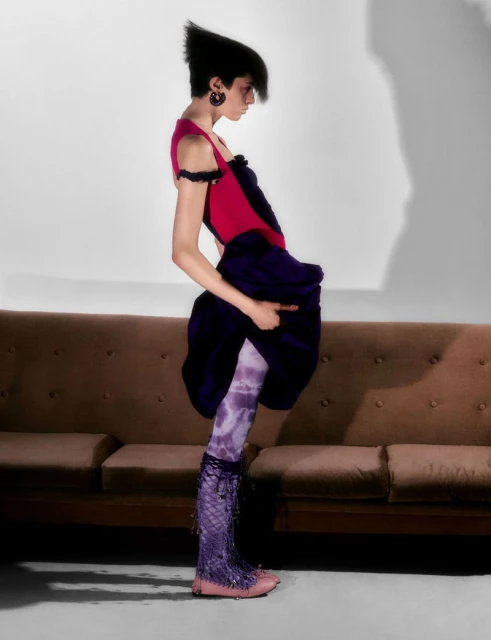 A woman in unique fashion stands in a photo studio with a brown couch, wearing purple patterned tights and dark purple skirt.