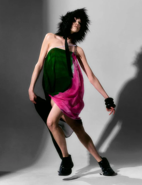 A fashionably dressed woman strikes a dynamic pose in a studio setting, showcasing a flowing green and pink dress with black accessories.