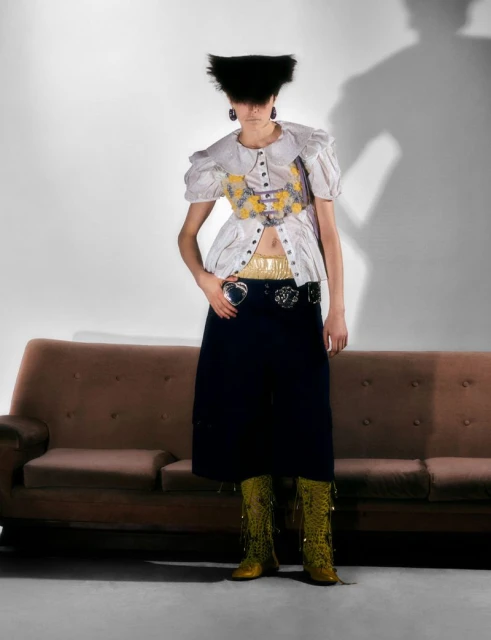 A woman with a unique hairstyle poses in a frilly white blouse, long dark skirt, and yellow boots, standing in front of a couch.