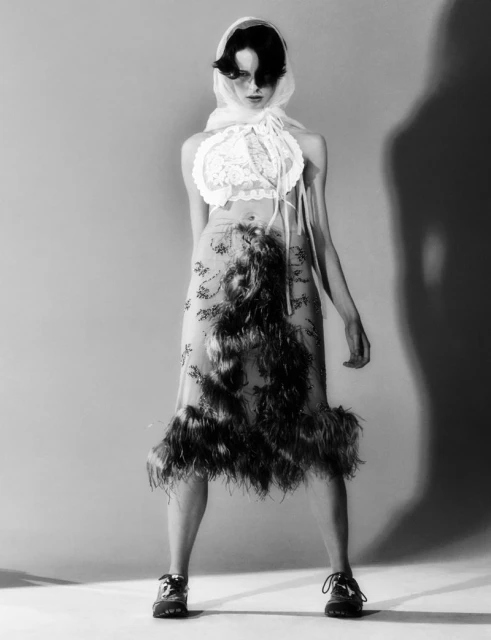 Monochrome fashion portrait of a woman wearing an ornate feather dress, headdress, and sneakers, posing in front of a light gray backdrop.