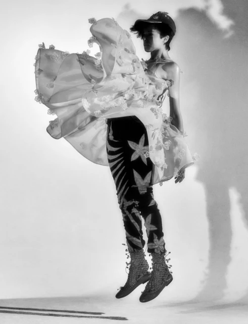 A person in patterned leggings and a semi-sheer, frilly dress leaps against a light background in a studio fashion shot.