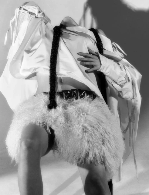Monochrome fashion shot of a model in an oversized white shirt, a fluffy skirt, a wide belt, and beaded straps, posing indoors with soft lighting.