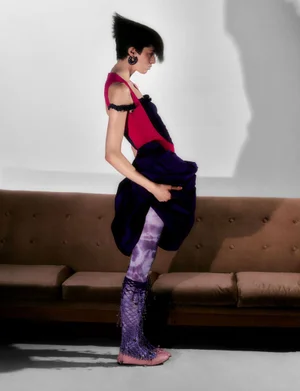 Model with styled dark hair and earrings standing in profile before a brown sofa wearing a vibrant dress and patterned tights