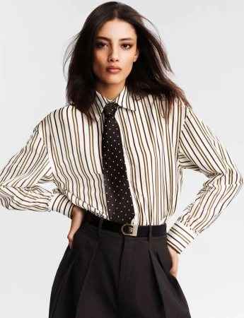 Model in a striped shirt and dark polka-dot tie posing against a white studio background for Heroine Magazine.
