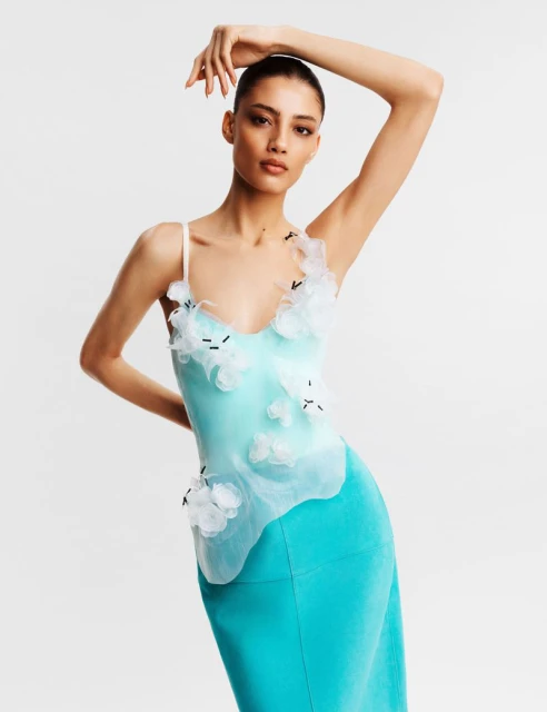 A woman in a light blue dress with white floral accents poses in a studio. She is facing forward with her right arm raised.
