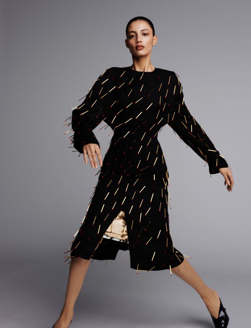A woman poses in a black and gold embellished dress, standing against a plain gray background in a fashion-style, eye-level studio shot.