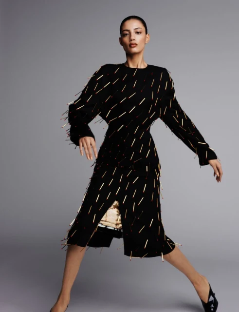 A woman poses in a black and gold embellished dress, standing against a plain gray background in a fashion-style, eye-level studio shot.