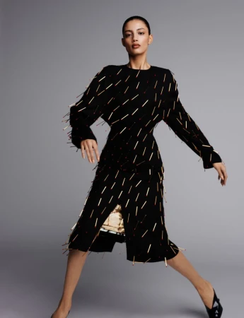 A woman poses in a black and gold embellished dress, standing against a plain gray background in a fashion-style, eye-level studio shot.