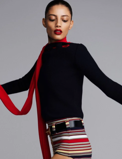 Fashion shot of a young adult woman in a black sweater and striped shorts, accessorized with a red scarf and black belt, against a grey background.