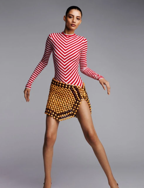 Fashion model poses in a red and white striped long-sleeve top and wood bead mini skirt against a grey backdrop, with studio lighting.
