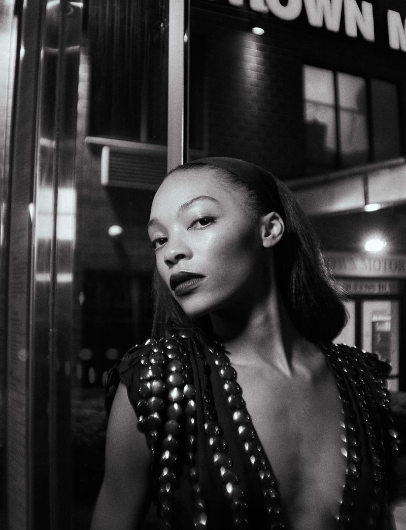 Monochrome portrait of a woman with dark lipstick and shoulder embellishments, posed in front of a dark building at night.