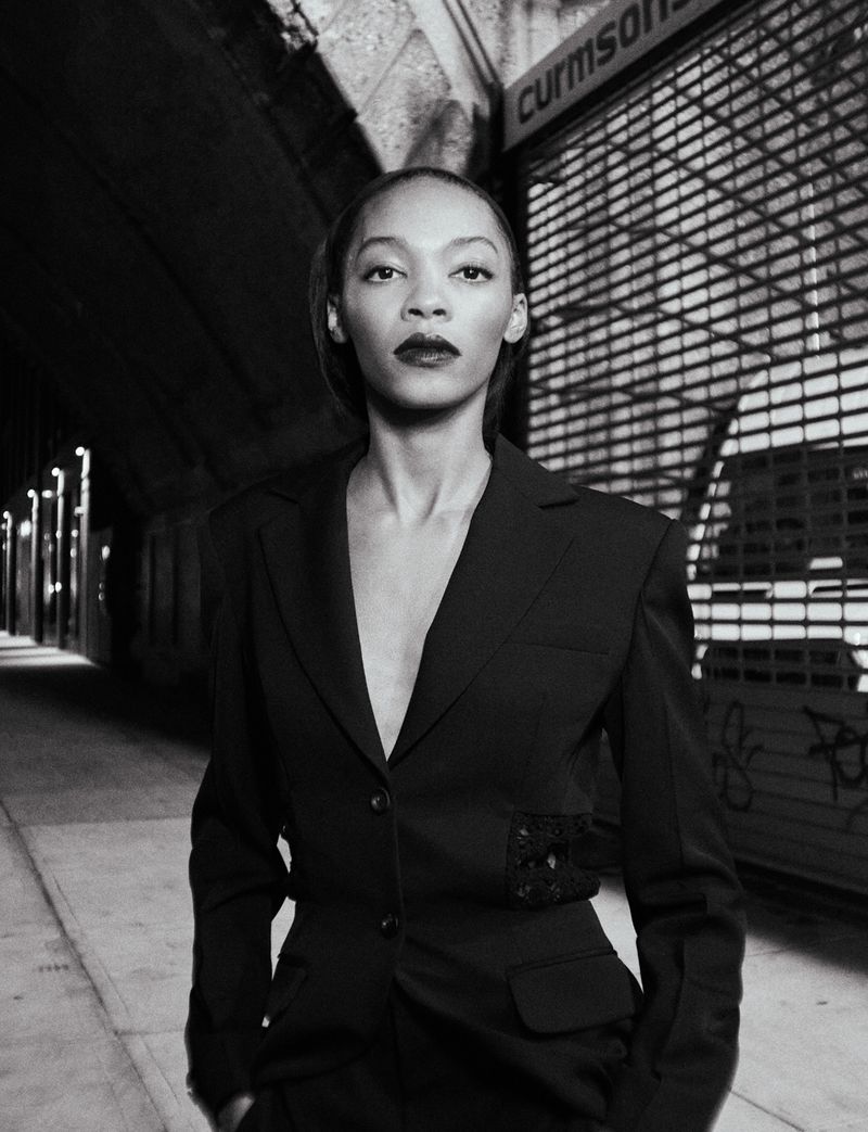 Black and white portrait of an adult woman in a dark business suit. The shot is set in an outdoor urban environment with concrete walls.