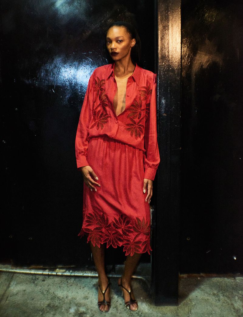 An adult female fashion model poses in an unbuttoned, reddish-pink dress with leafy embroidery, paired with strappy heels against a dark background.