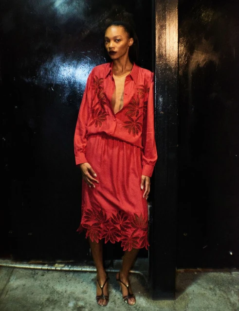 An adult female fashion model poses in an unbuttoned, reddish-pink dress with leafy embroidery, paired with strappy heels against a dark background.