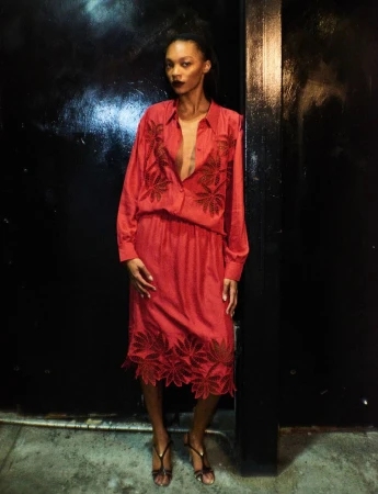An adult female fashion model poses in an unbuttoned, reddish-pink dress with leafy embroidery, paired with strappy heels against a dark background.