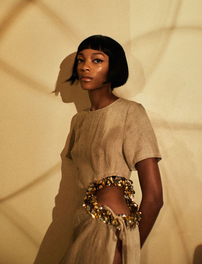 Elegant, dark-skinned woman in a beige dress with a jeweled cutout, posing indoors with geometric shadow patterns on the neutral wall.