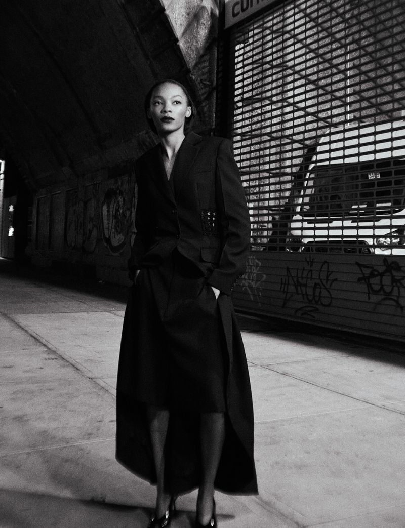 A woman in a blazer and skirt stands in an urban setting with dark lighting, showcasing fashion with street style.