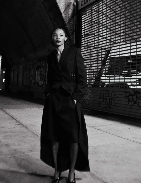A woman in a blazer and skirt stands in an urban setting with dark lighting, showcasing fashion with street style.