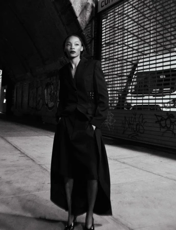 A woman in a blazer and skirt stands in an urban setting with dark lighting, showcasing fashion with street style.