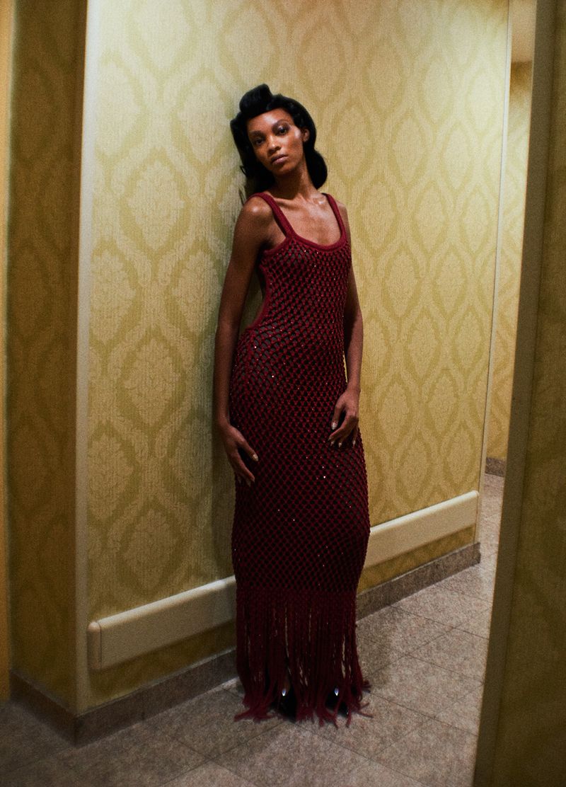 An adult woman with dark hair and skin poses in a burgundy beaded dress in a hallway with patterned wallpaper and light-colored tile flooring.