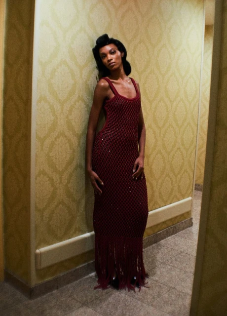 An adult woman with dark hair and skin poses in a burgundy beaded dress in a hallway with patterned wallpaper and light-colored tile flooring.