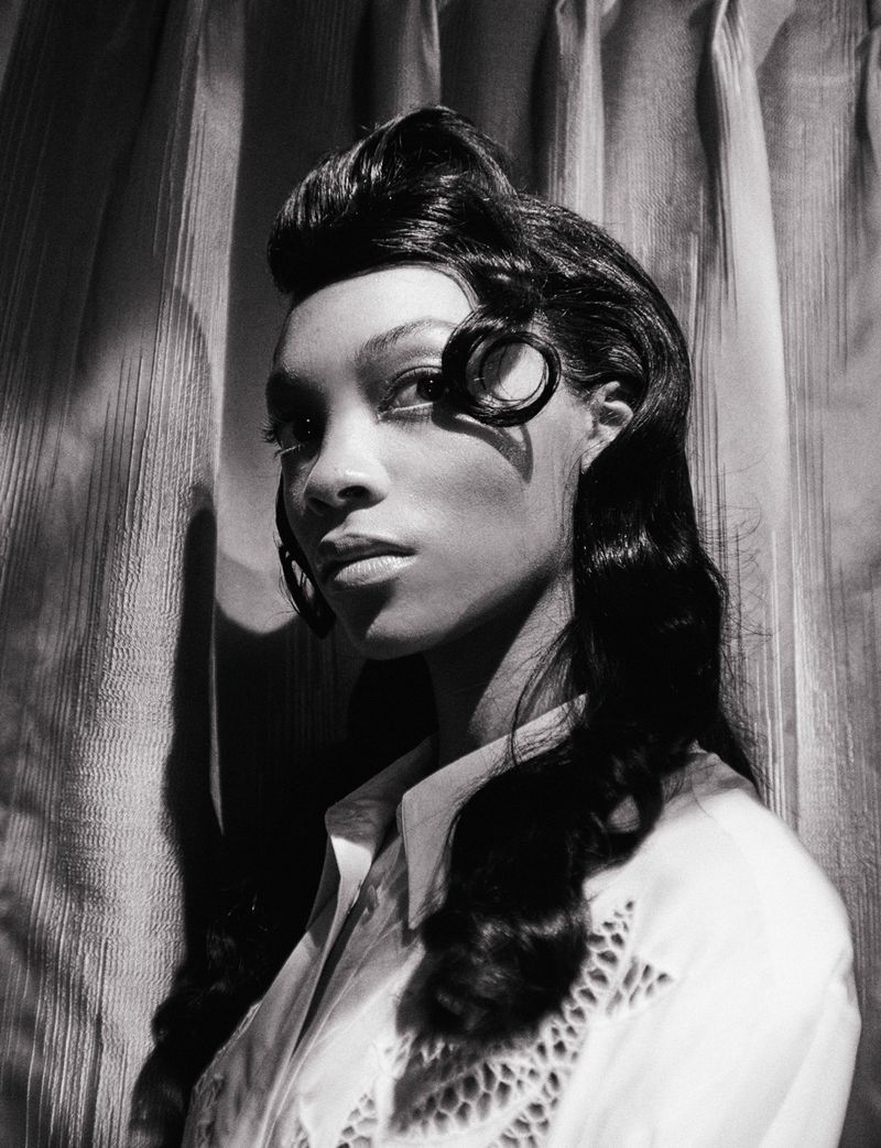 Black and white portrait of an African American woman with curled dark hair and a white, collared, eyelet shirt against a textured curtain.