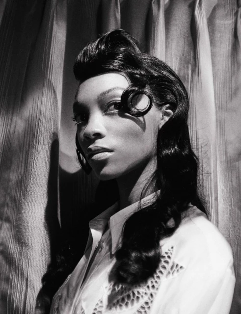 Black and white portrait of an African American woman with curled dark hair and a white, collared, eyelet shirt against a textured curtain.