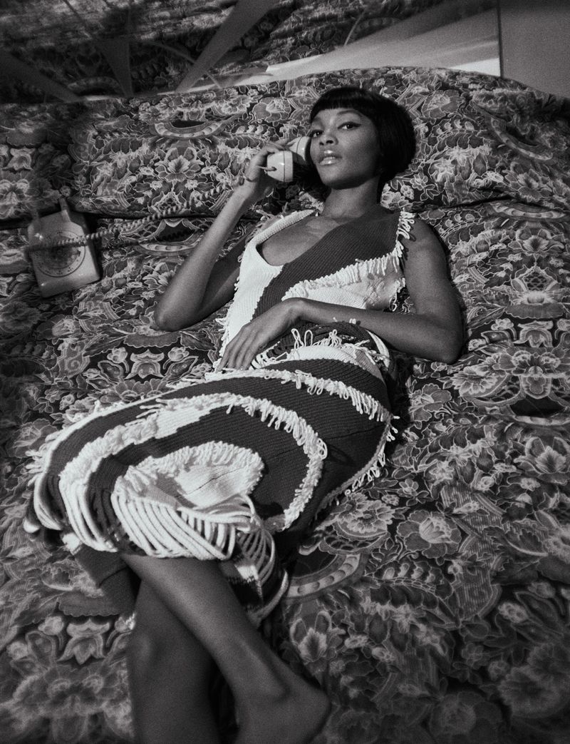 Black and white portrait of a woman in a fringed dress lying on a patterned bed while talking on a vintage telephone receiver.