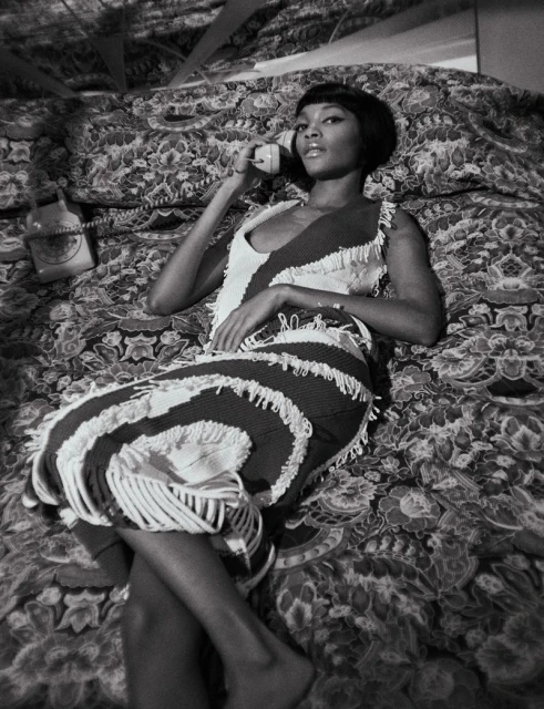 Black and white portrait of a woman in a fringed dress lying on a patterned bed while talking on a vintage telephone receiver.