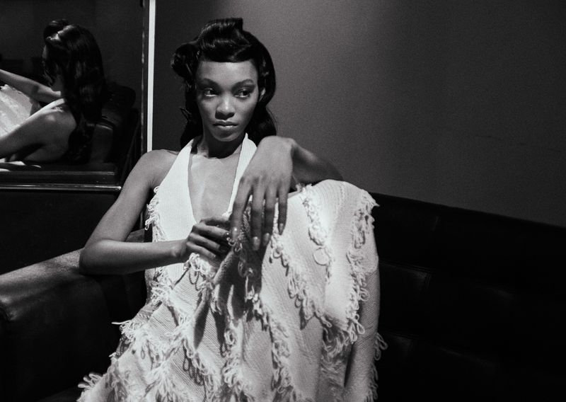Black and white portrait of a woman in a textured fringed halter dress seated indoors, reflected in a mirror.
