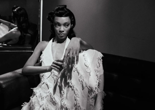 Black and white portrait of a woman in a textured fringed halter dress seated indoors, reflected in a mirror.