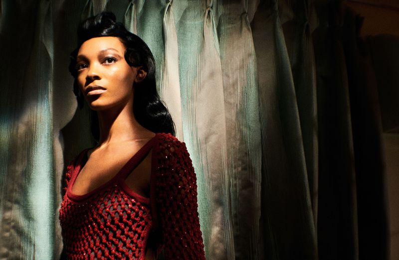Portrait of an African American woman with styled black hair, wearing a maroon knit top, posing indoors against draped textured curtains.