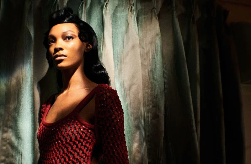 Portrait of an African American woman with styled black hair, wearing a maroon knit top, posing indoors against draped textured curtains.
