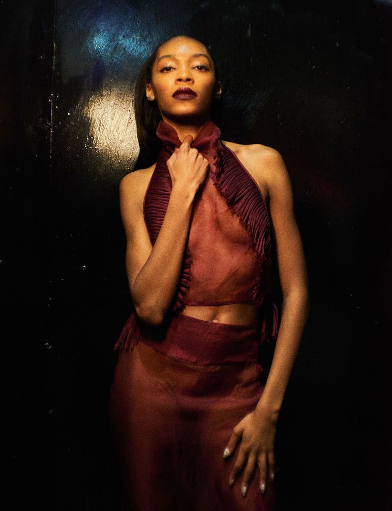 Elegant woman in a maroon two-piece outfit poses in front of a dark background, hands near her neck, with a confident expression.