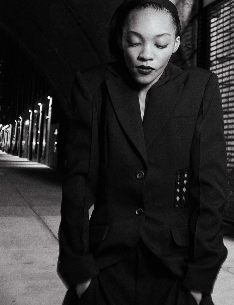 Black and white portrait of an African-American woman in a dark blazer and pants, with a dark background suggesting a urban setting.