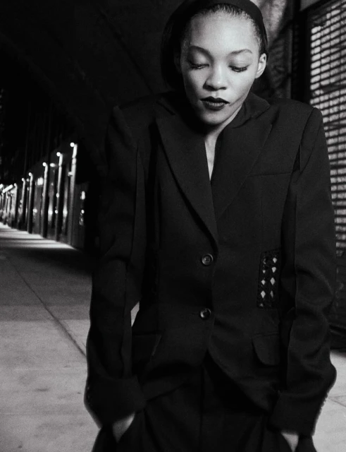 Black and white portrait of an African-American woman in a dark blazer and pants, with a dark background suggesting a urban setting.