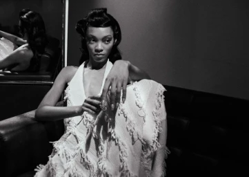 Black and white portrait of a woman in a textured fringed halter dress seated indoors, reflected in a mirror.