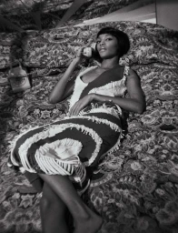 Black and white editorial portrait of a woman lying on a patterned bed holding a vintage telephone receiver to her ear