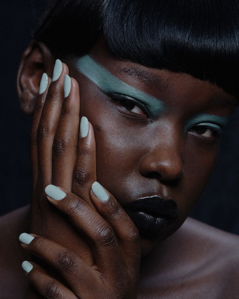 Close-up portrait of a Black woman with green eyeshadow and pale green nails against a dark background in a beauty style image.