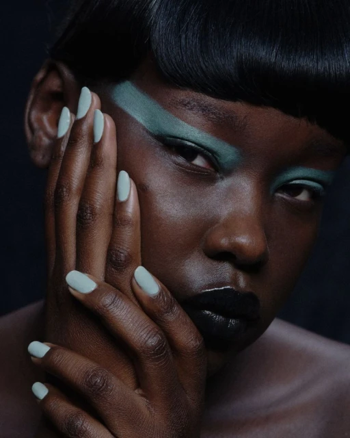 Close-up portrait of a Black woman with green eyeshadow and pale green nails against a dark background in a beauty style image.