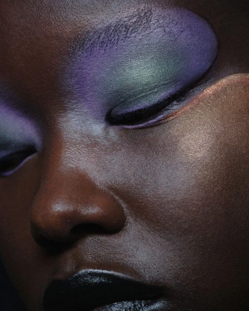 Close-up portrait of a woman with closed eyes and artistic makeup featuring purple and green eyeshadow and dark lipstick.