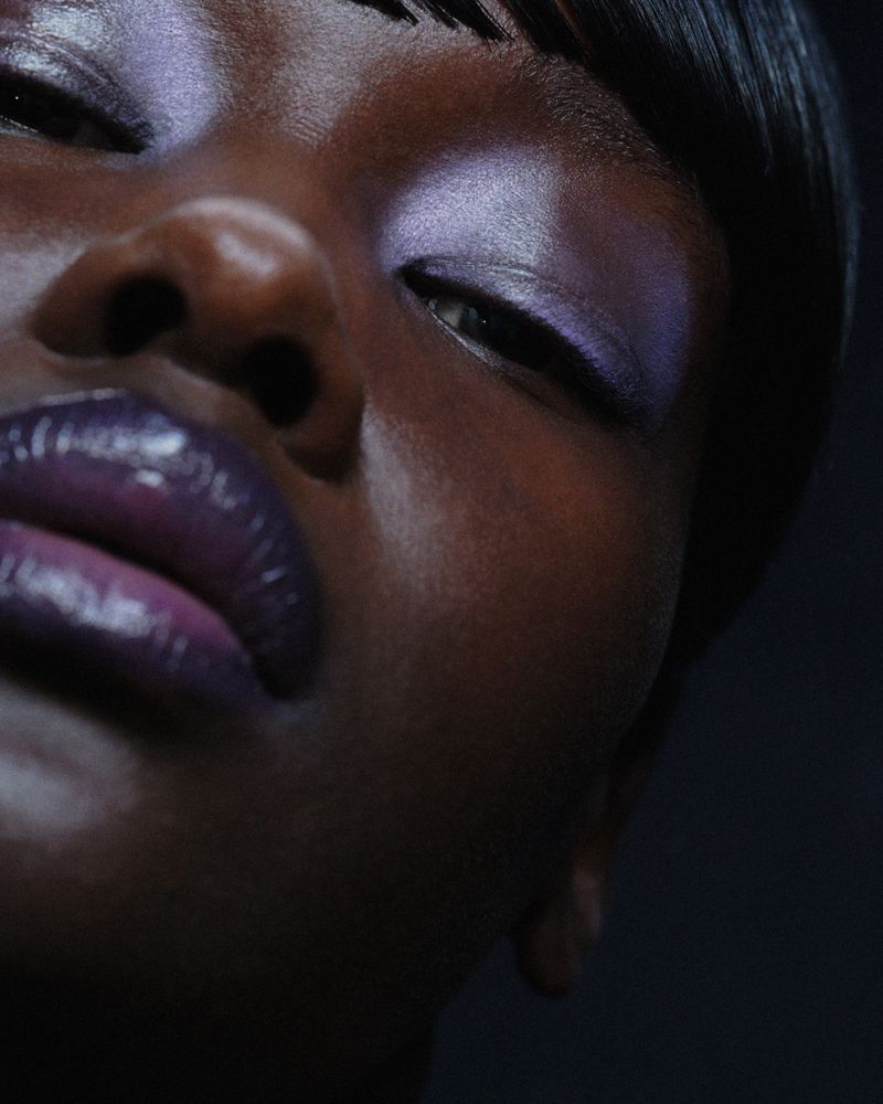 Close-up portrait of an African American female with dark, glossy skin and a beauty makeup look featuring iridescent eyeshadow and glossy lips.