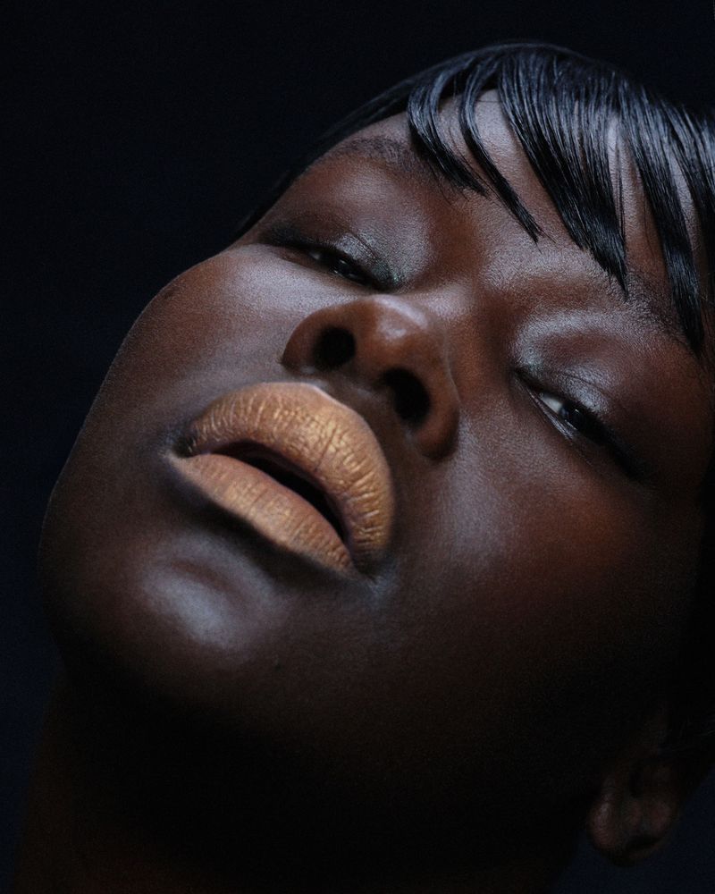 Close-up portrait of a Black woman with glossy lips and short dark hair against a dark backdrop in a beauty and fashion setting.