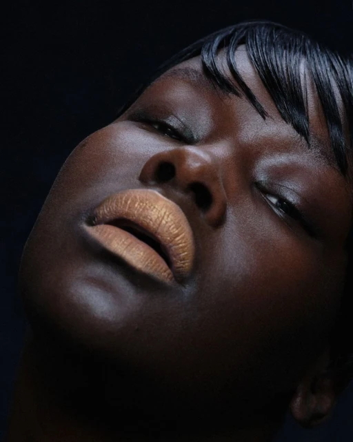 Close-up portrait of a Black woman with glossy lips and short dark hair against a dark backdrop in a beauty and fashion setting.