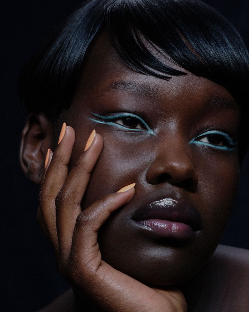 Close-up beauty portrait of a Black model with blue winged eyeliner and peach-colored manicured nails against a dark background.
