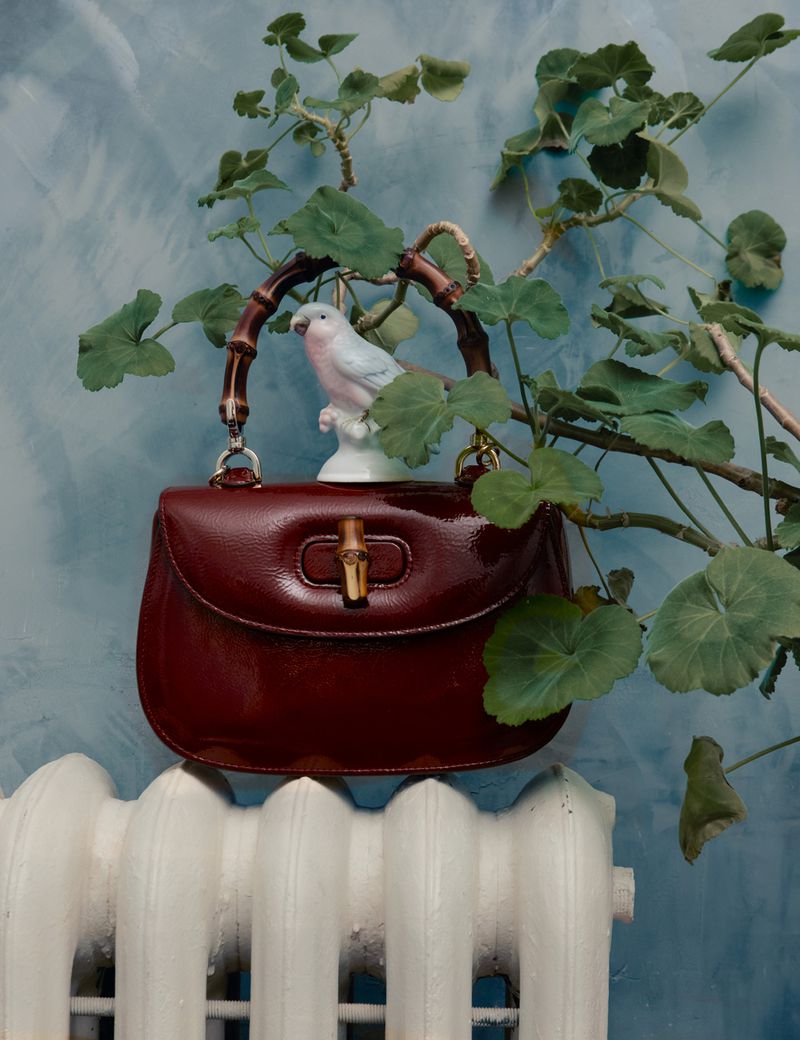 Burgundy leather Gucci handbag with bamboo handle, ceramic bird figurine, and potted plant against a blue textured wall and radiator.